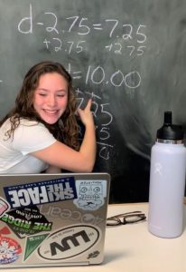 student tutor smiling looking at computer and pointing to chalkboard with a math problem