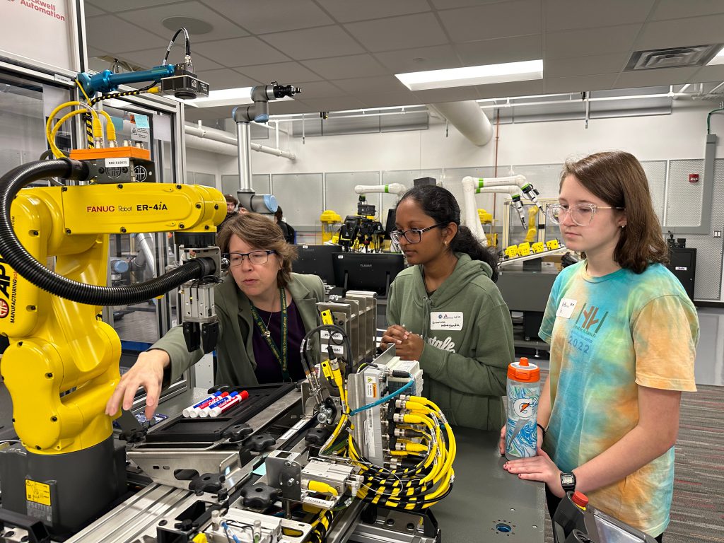 Two students look on as a professional demonstrates some of the manufacturing equipment.