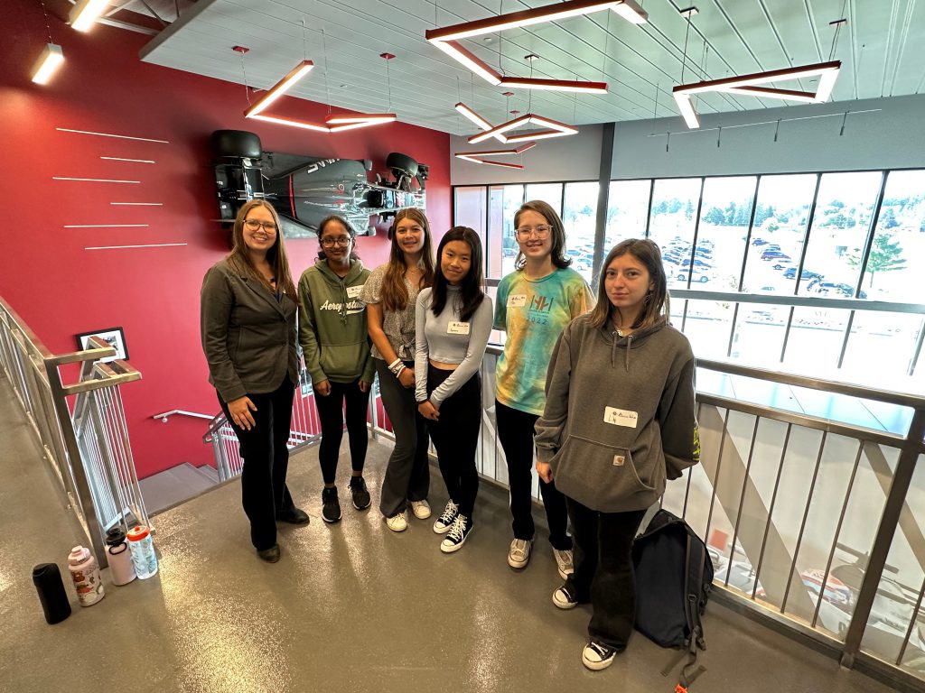 The five Shaker students and their teacher pose together in front of a race car mounted on the wall.