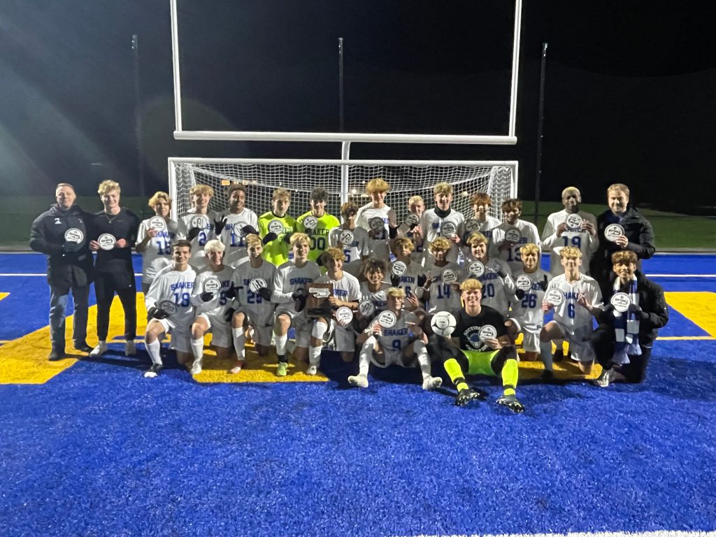An image of the sectional champion Shaker boys varsity soccer team posing with their championship patches after the win.